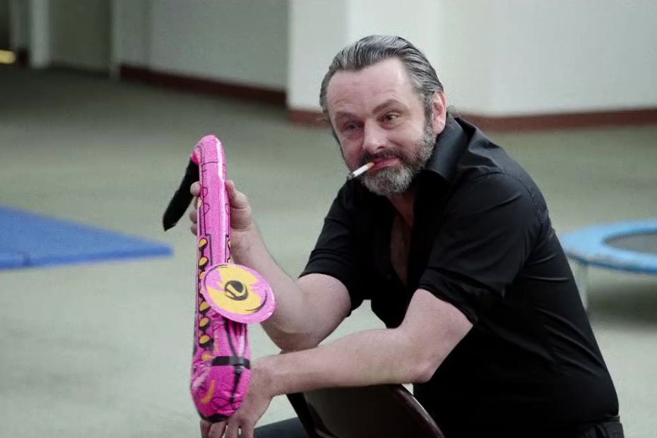 Michael Sheen as Carl Flossy, dance coach. He’s holding an inflatable pink saxophone 🎷 