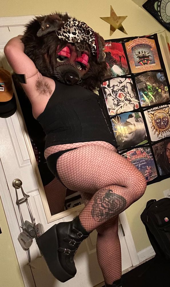 Bailey (brown mastiff mask) standing facing right with one leg kicked up and her arms behind her head. She has on 5in black platform boots, fishnets, panties, bra and corset with a wine colored lace up back. She is showing off her hairy armpit and all her fancy jewelry; a mix of bronze, silver and gold