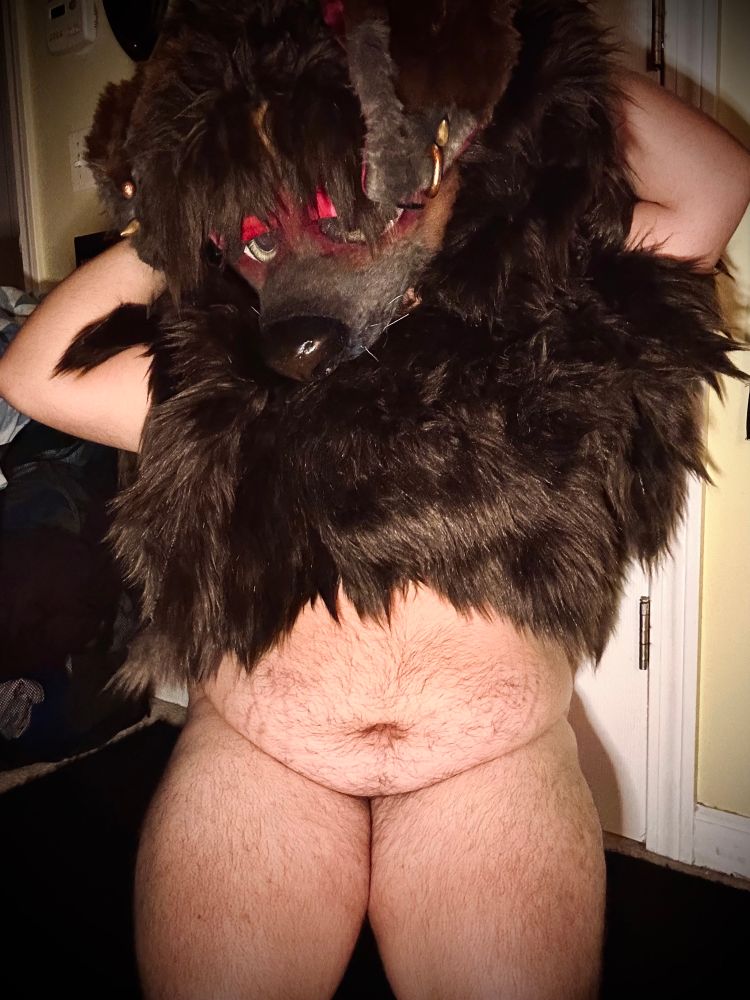 Bailey (fur poncho and mask) kneeling in front of the camera with her arms behind her head, looking out from under her bangs at the camera