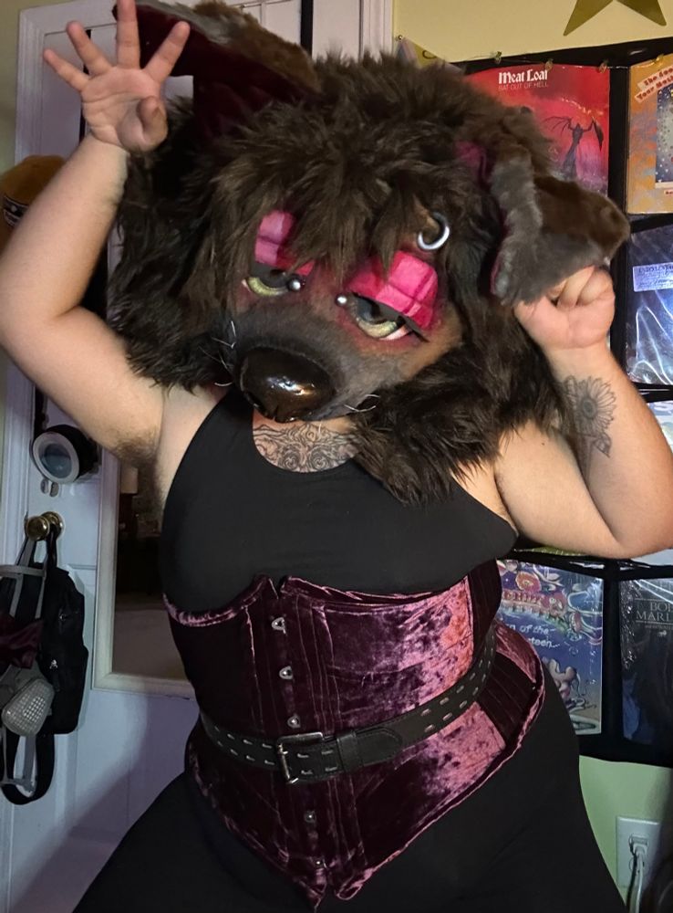 Bailey (brown mastiff mask) in a stuffed black bra, leggings and a burgundy corset with a black belt with silver hardware. She has a few silver piercings too, most notably between her eyes and his eyebrow ring. He is slightly squatting making her hips look wide and her waist cinched with both arms up playing with his ears. He looks into the camera with big green half-lidded eyes