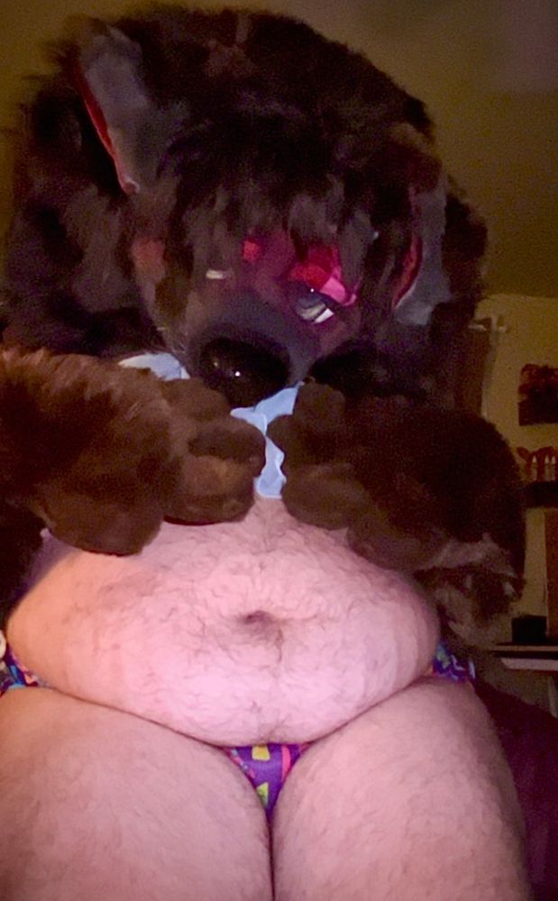 Bailey holding her chest in her paws with her head bowed slightly down, looking up from under her heady eyelids and lashes. Her legs are pressed together but the chub on his fupa and love handles is poking out with the funky purple boot fabric of her panties