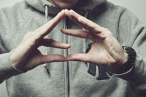 Photo couleurs format paysage. Gros plan sur des mains blanches formant la lettre S en joignant les pouces, les index au centre et le reste des doigts joints aussi. Derrière une personne avec sweat gris.