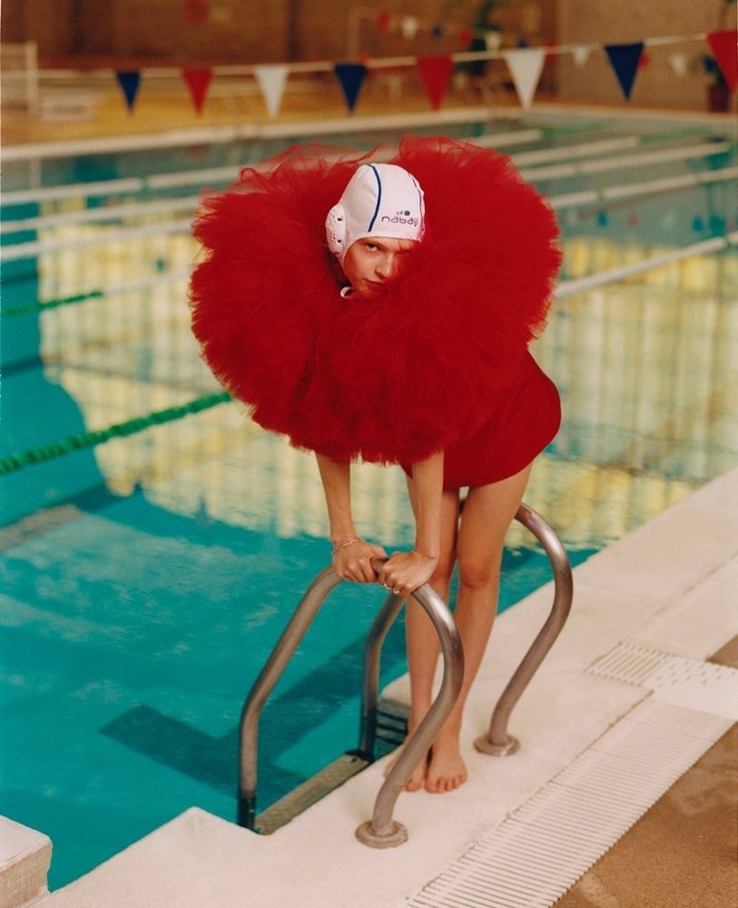 Photo couleurs format portrait.
A l'intérieur d'une piscine publique une femme blanche se tient debout sur le rebord entre les bras d'une échelle en métal qui sort de l'eau, cadrée en plein pied. Elle se tient les mains appuyé sur un des bras de l'échelle. Elle est habillée avec une tenue rouge qui laisse voir ses avant-bras et ses jambes et pieds nus. Sur le haut du corps la tenue est un énorme froufrou recouvrant tout le buste et une partie du visage. Sur la tête un bonnet de bain blanc recouvre les cheveux.