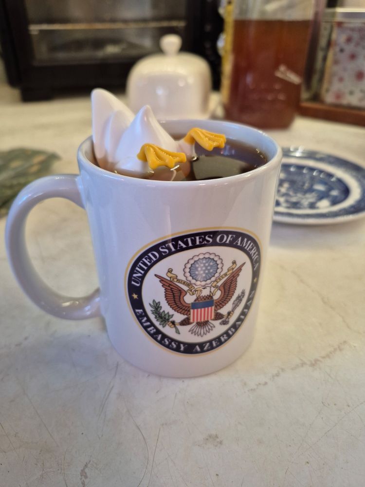 A white mug with a tea infuser shaped like a duck's butt floating in it. The mug is of a US seal that says "United states of America Embassy Azerbaijan".