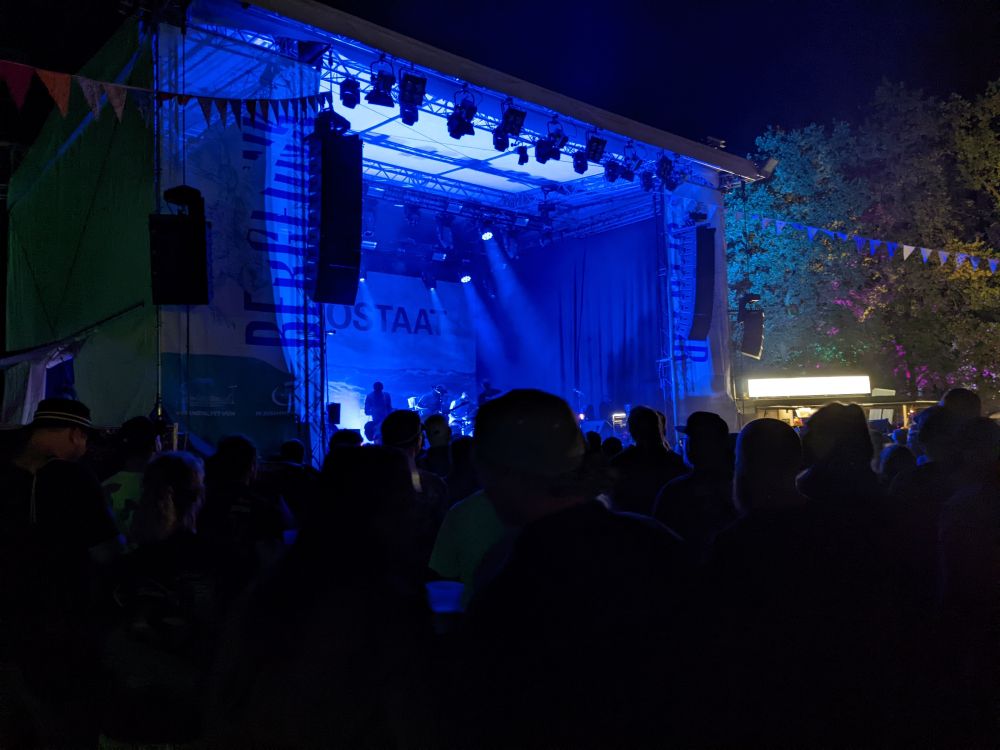 Menschen von hinten vor einer blau beleuchteten Bühne im Dunkeln. An der Bühne ein Banner mit Bergfunk (das Festival) und Turbostaat (die Band die gerade spielt)