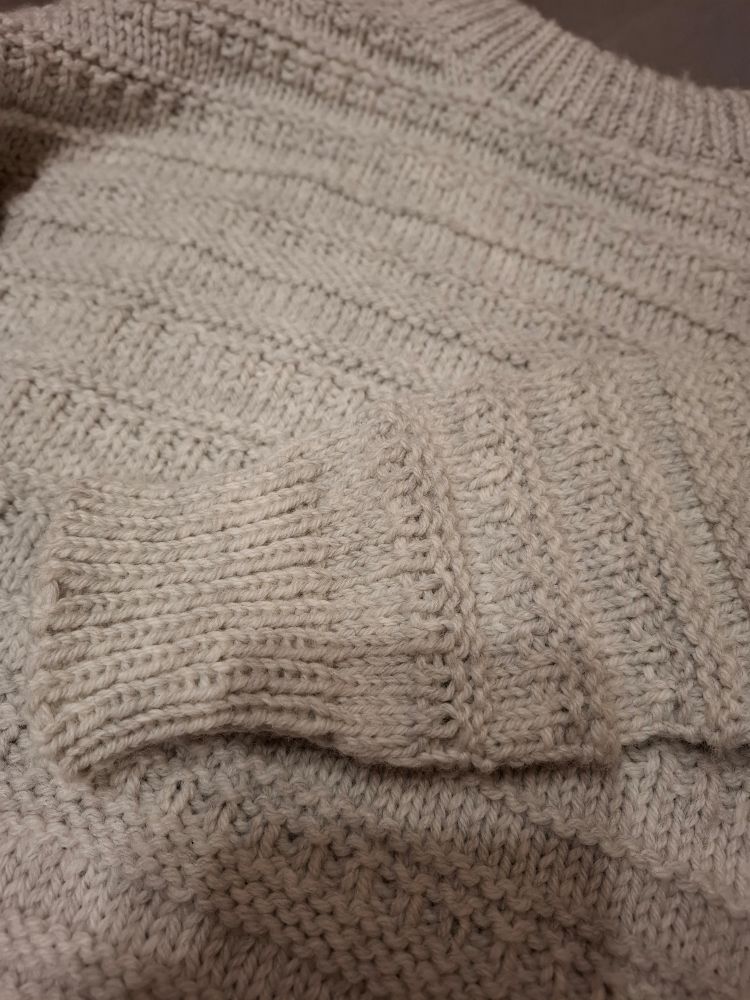 Nahaufnahme vom Ärmel mit Rippbündchen und Vorderteil. 
Beispiele für das Muster:
Mehrere Reihen glatt rechts, unterbrochen von einer oder mehrerer Reihen linker Maschen, dann zwei Reihen abwechselnd links/rechts, was auch eine Art geripptes Muster ergibt. Diese Art wiederholt sich in unterschiedlicher Kombination auf dem gesamten Pullover. 