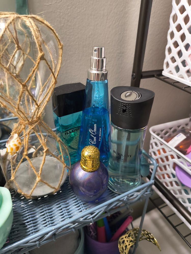 A blue metal shelf holds four bottles of perfume and cologne, an hourglass with twine, and a white plastic basket with various small items in the background.