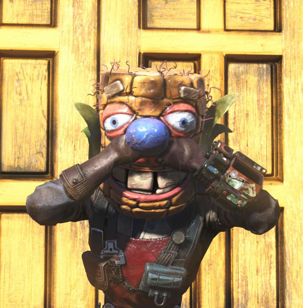 A character in Fallout 76 wearing a corn cob mask with large eyes, a blue nose, and big teeth, standing in front of a yellow wooden door.