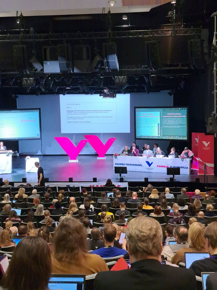 Sali, jossa paljon ihmisiä, lavalla puheenjohtajisto.