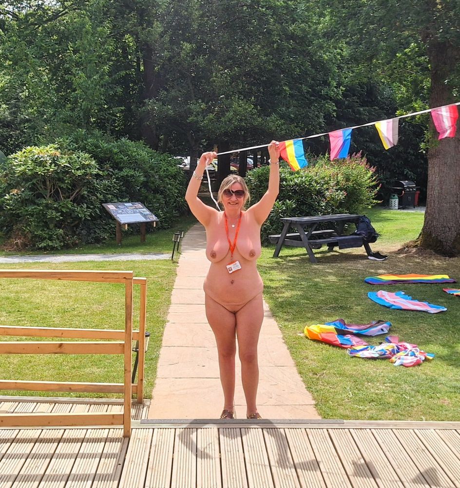 Helen Berriman stand nude smiling to the camera and holding the end of some gay pride bunting above her head. She is on a path in front of some decking with green hedges behind. It's a sunny day.