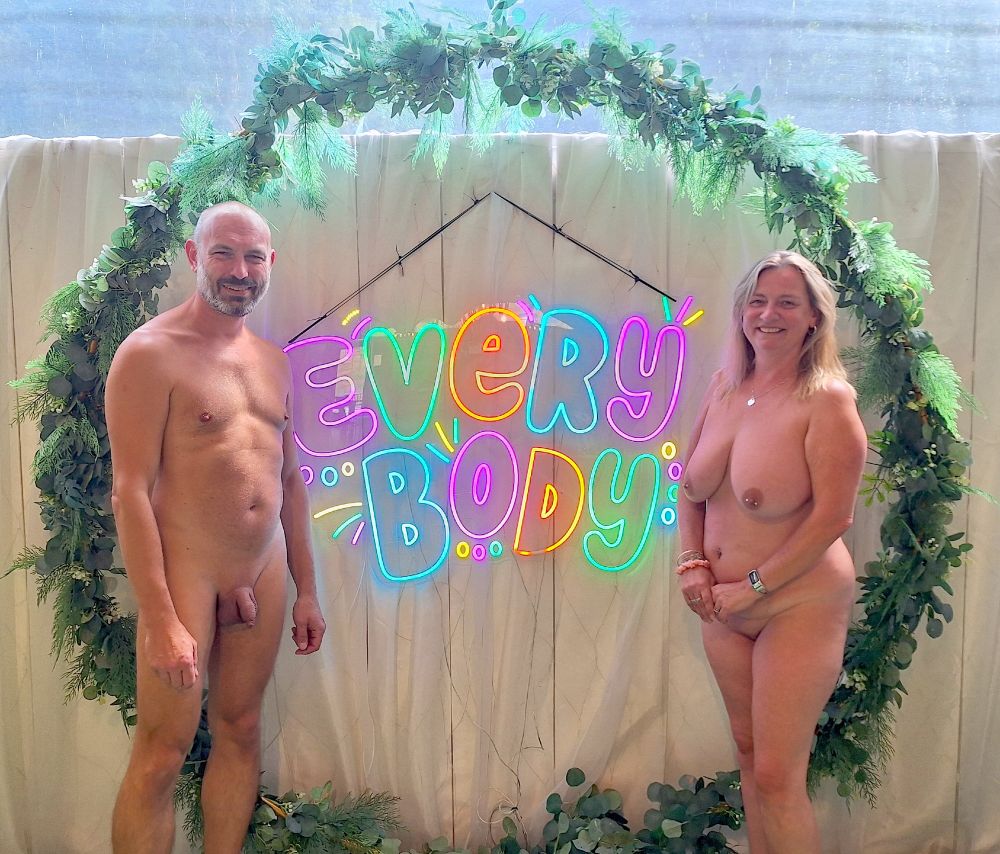 A man and a woman stand either side of a multi-coloured light-up sign that reads "Every Body". A green wreath a fake leaves surrounds them both and the sign.