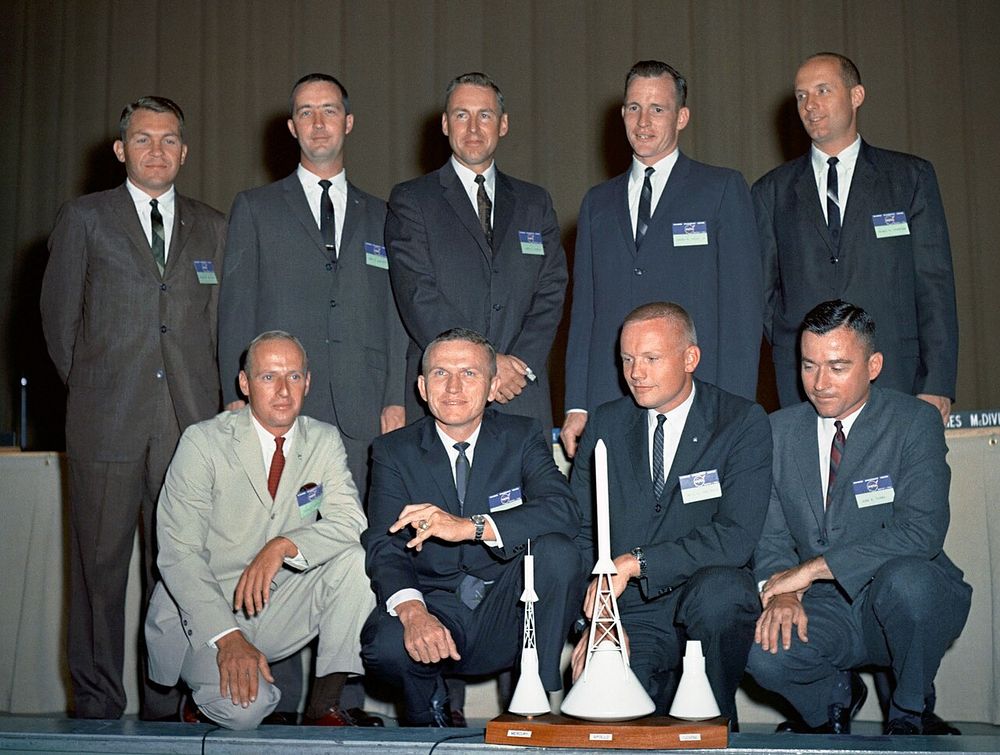Colour photo of nine astronauts selected in 1962, all in suits and ties with model spacecraft in front of them. In the back row, standing, are Elliot See, James McDivitt, James Lovell, Edward White and Thomas Stafford. Crouching in front are Charles 'Pete' Conrad, Frank Borman, Neil Armstrong and John Young.