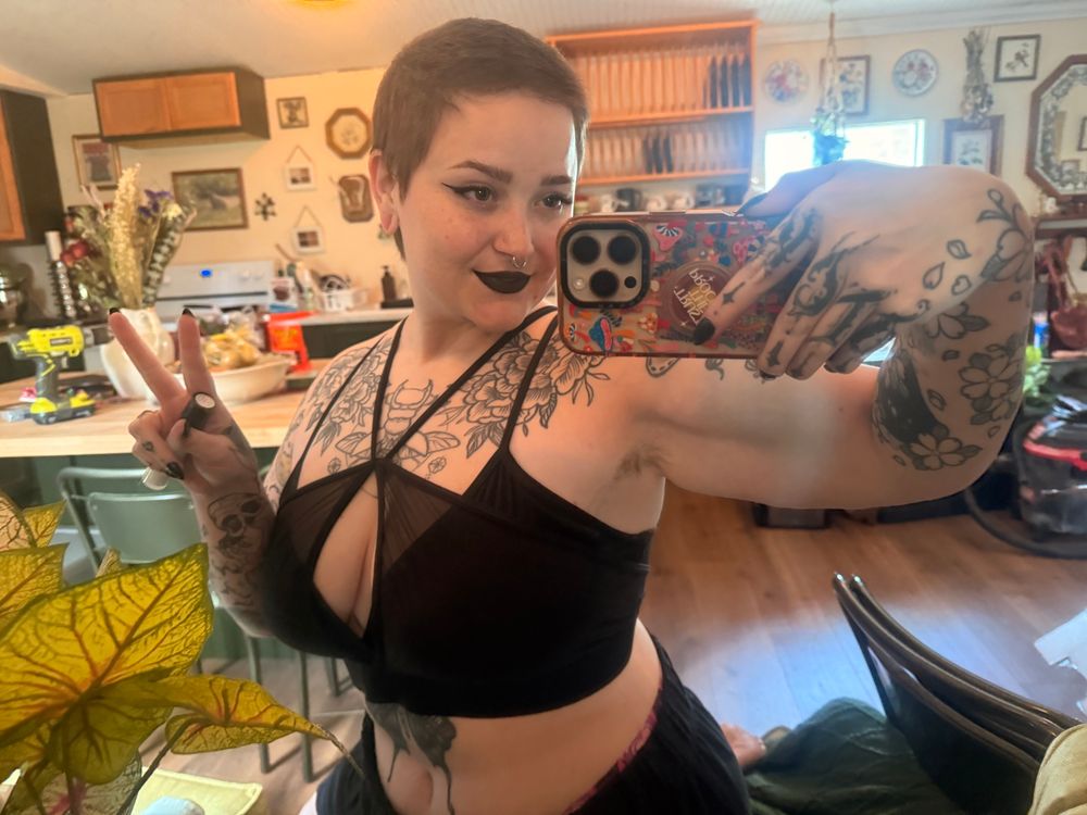 mirror selfie of a person in an eclectic home with black lipstick and a black bralette.