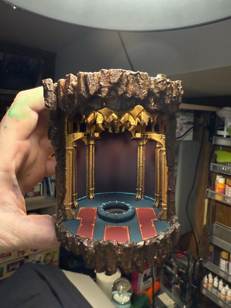 photograph of a miniature version of the room the Mammon Machine is suspended in from Chrono Trigger being held by a hand with a messy workspace out of focus in background. Room is circular and grand looking about 5” tall and 3” in diameter