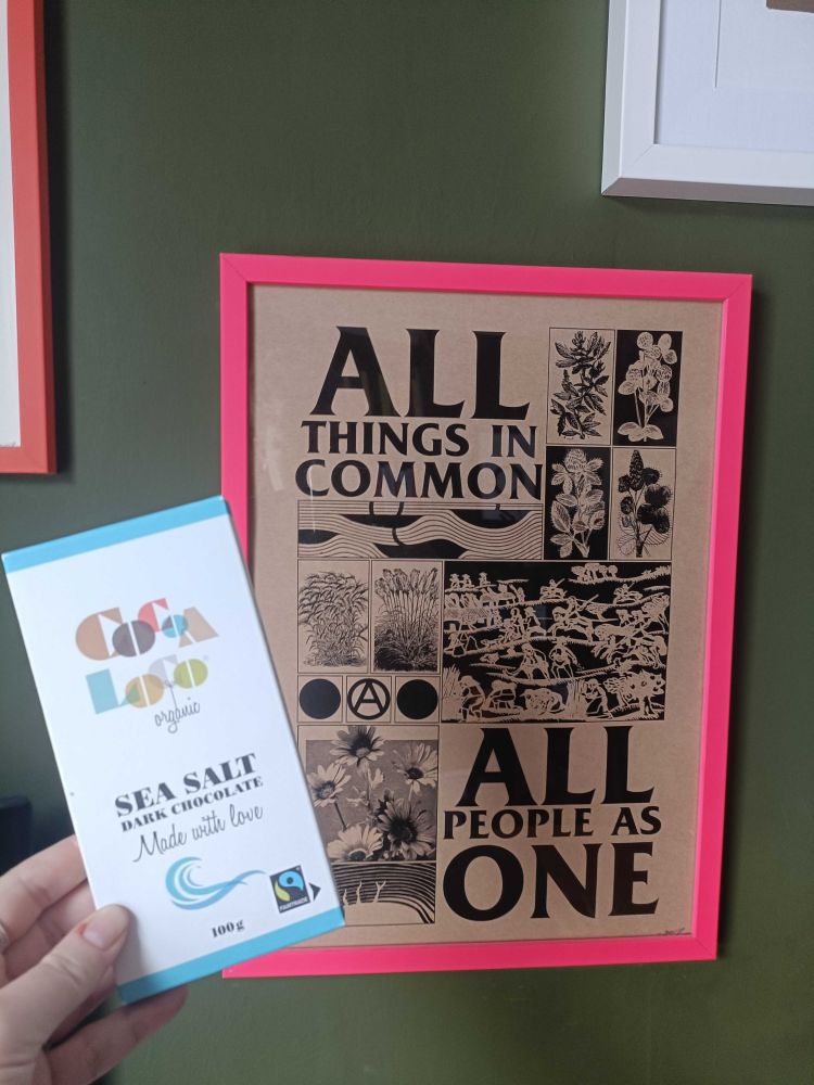Ellie holds up her chocolate bar gift with some artwork in the background which reads: All things in common, all people as one.

