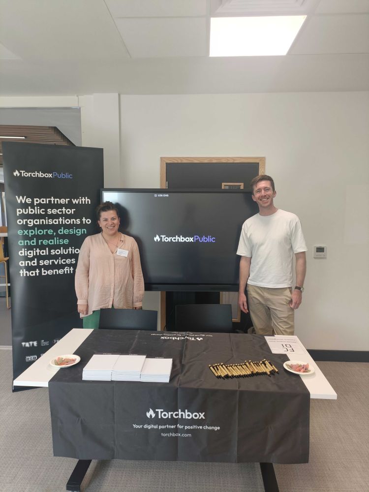 Ellie and Mark stand behind our branded table at Local Gov Camp - our roller banner reads: We partner with public sector organisations to explore, design and realise digital solutions and services that benefit all. 