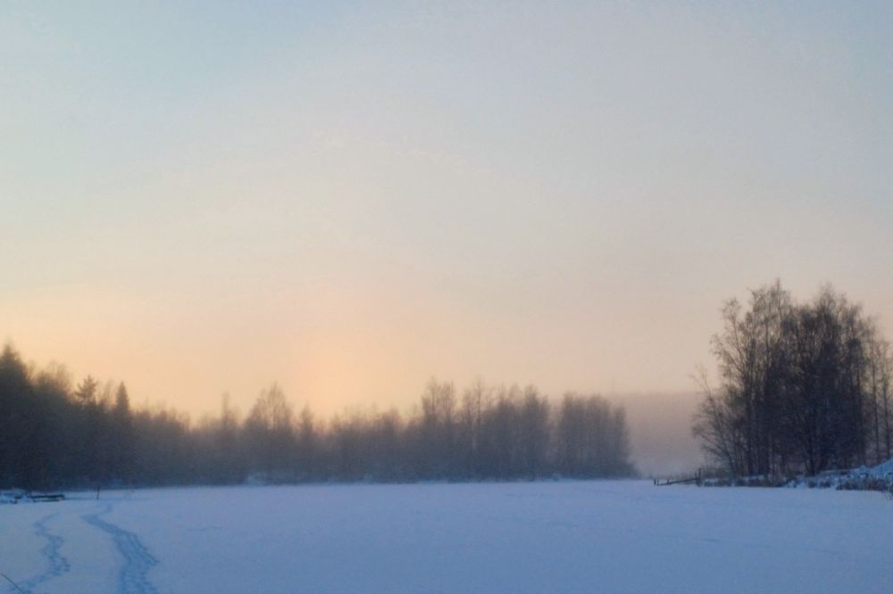 Taivaalla leijuva lumipöly hehkuu lämpimän oranssina viileän sinisävyisen järven jään yllä.