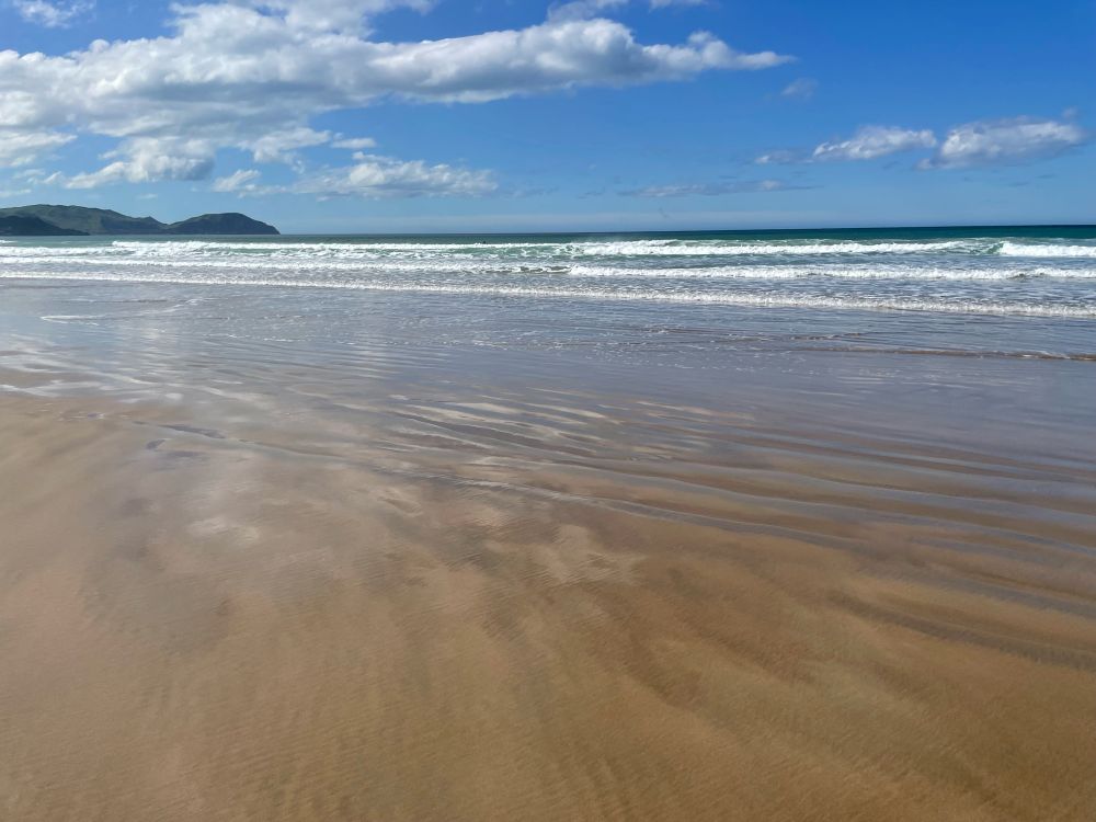 On a sandy beach looking. out at the ocean with scattered fluffy clouds and blue skies.  Some green hills in the distance on the left.