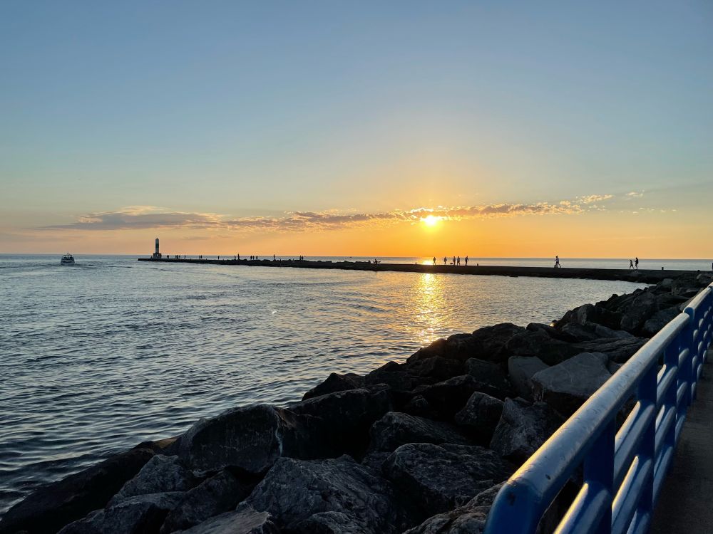 Standing on the beginning of a pier looking out towards the sun setting over Lake Michigan with a thin line of clouds stretching across most of the horizon. 