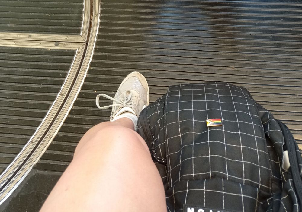 Bild meines Beins, Rücksack und Straßenbahn Fußboden. An meinem Rücksack ist ein rechteckiger Pin mit der Progress pride flag befestigt.