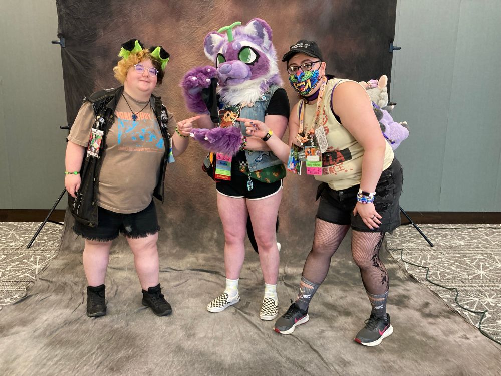 Photo of left to right, Scamper, Joy from Troy fursuit, and Artie in front of a photo backdrop with Joy holding a sock and Scamper and Artie pointing at it