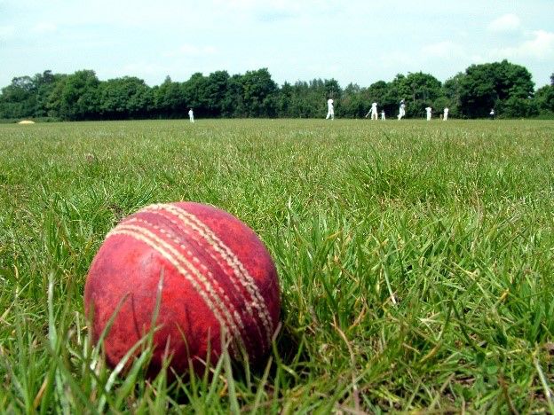 A red cricket ball on grass, with players in whites visible in the background.