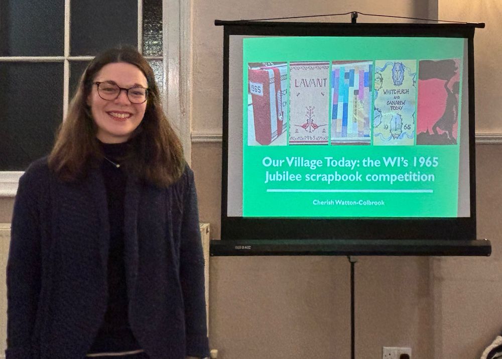 Cherish, standing in front of a slide titled ‘Our Village Today: the WI’s 1965 Jubilee scrapbook competition’ by Dr Cherish Watton-Colbrook. The slide shows the front covers of several scrapbooks.