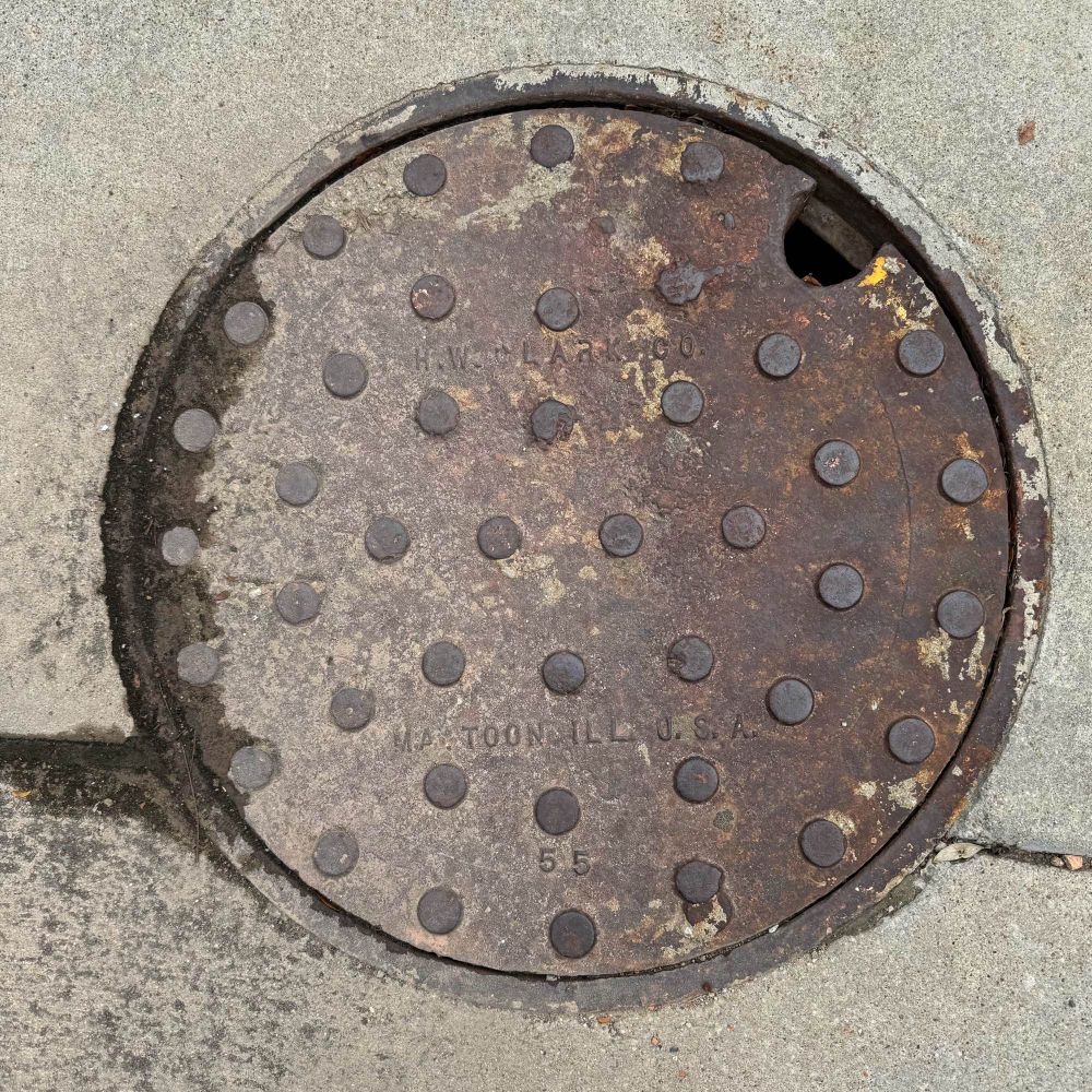 Round iron utility cover with a pattern of round raised bumps and the words “H.W. CLARK Co. MATTOON, ILL, U.S.A. 55”