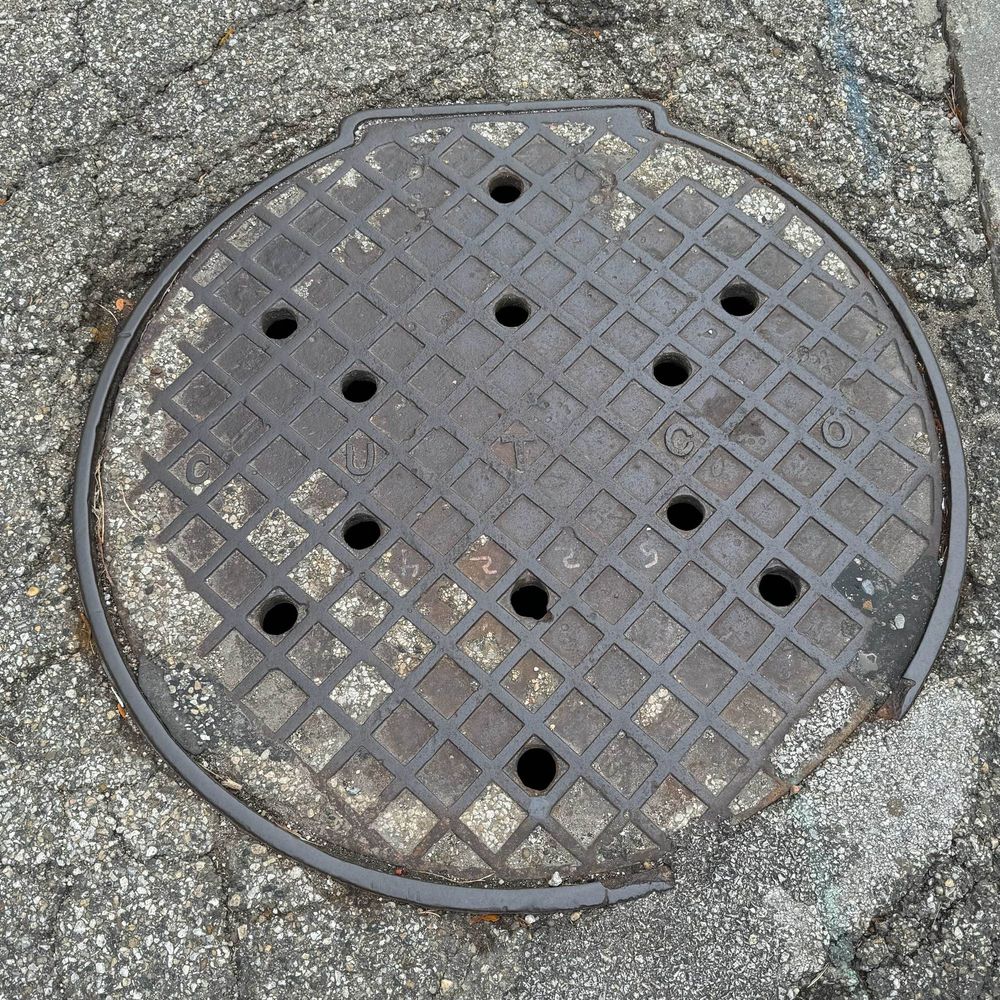 Round iron utility cover with a smattering of round holes in it and a crosshatch pattern, plus the letters C U T C O