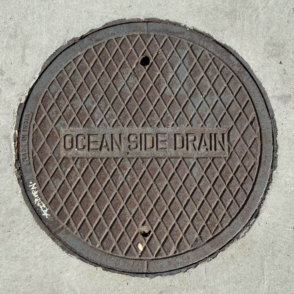 Round iron cover with a diamond grid pattern and the words “Ocean Side Drain” in a box in the center.