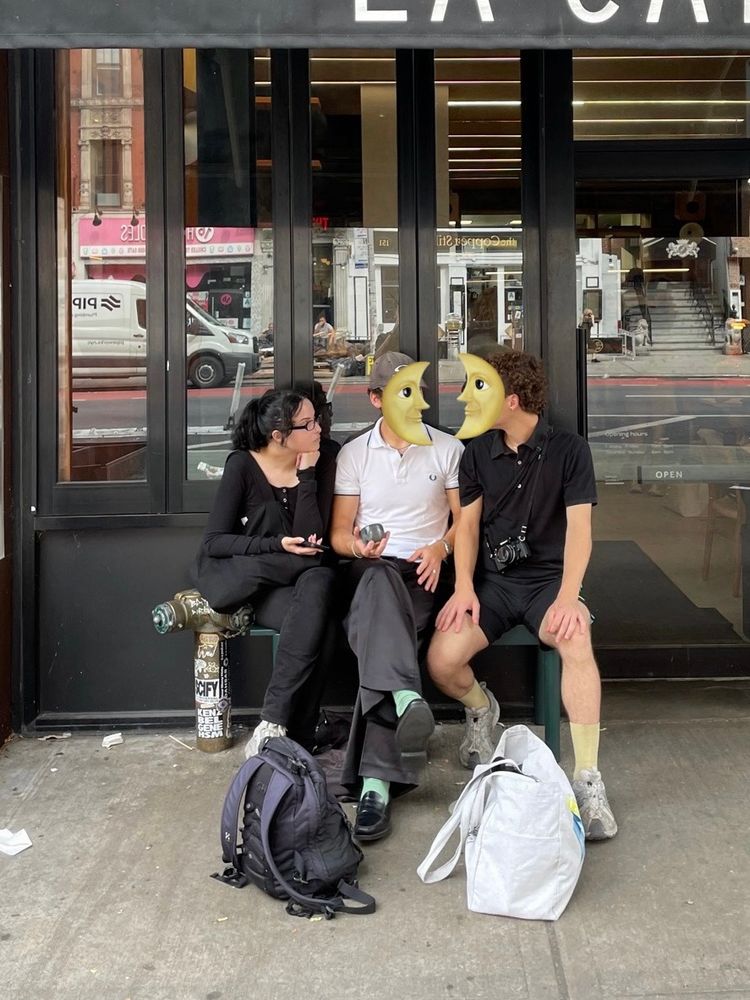 Three people in their twenties sitting on a small bench in front of a coffee shop with a black facade and tall narrow windows.
The first from the left is a woman resting her head on her fist and seemingly listening intently to the conversation going on between two men sitting next to her.