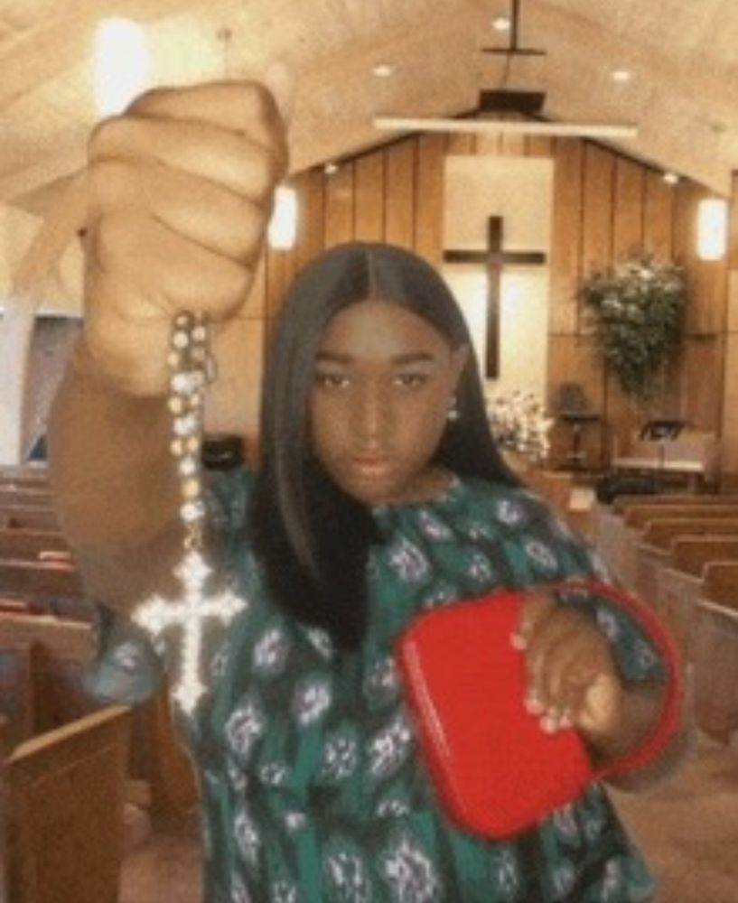 Woman in a church holding a rosary out in front of her, towards the camera, with one hand, and clutching a red handbag to her chest with the other