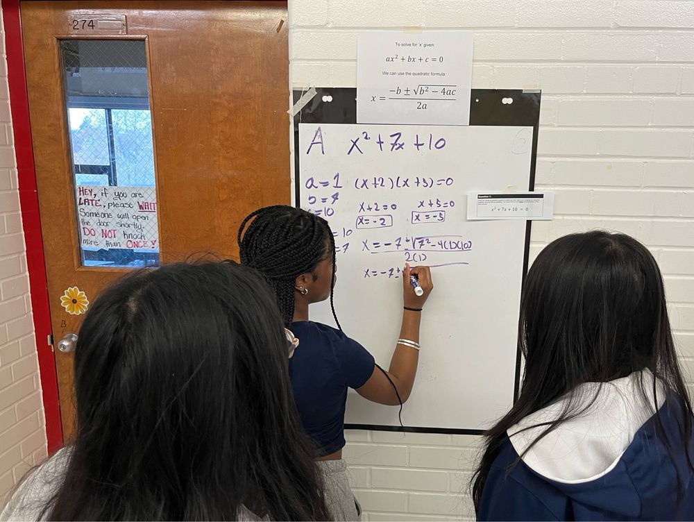 Three female students working in front of a whiteboard. One student is writing and using the quadratic formula.