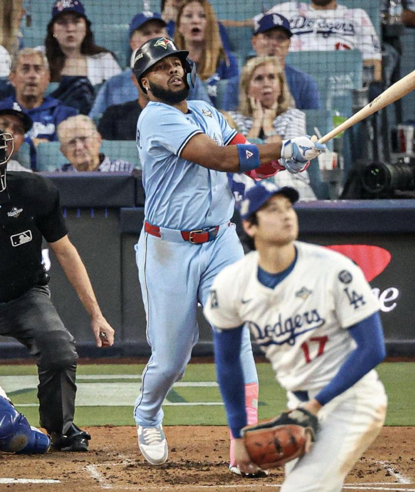 A wonderful shot of Vladimir Guerrero Jr. admiring his pulled home run to left field yesterday off Shohei Ohtani. Guerrero is a very stocky Black man of dark complexion with a heavy black beard. He is dressed in a gorgeous powder blue road uniform; his arms are extended where he has completed his swing. He is looking up to the right of the frame (left field) and Ohtani, a tall handsome Japanese pitcher, is in the foreground, turned around and looking to the same place. The crowd in the front row seats behind (maddeningly empty as always at Dodger stadium) are all looking, some in horror and some in concern, in the same direction. 