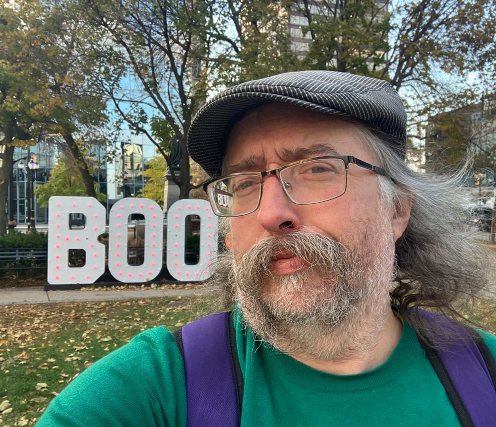 I am standing in Gore Park. I am swearing a blue hat and green sweater and I have a very skeptical look on my face. Beside me are very large letters with chasing lights all over that say BOO