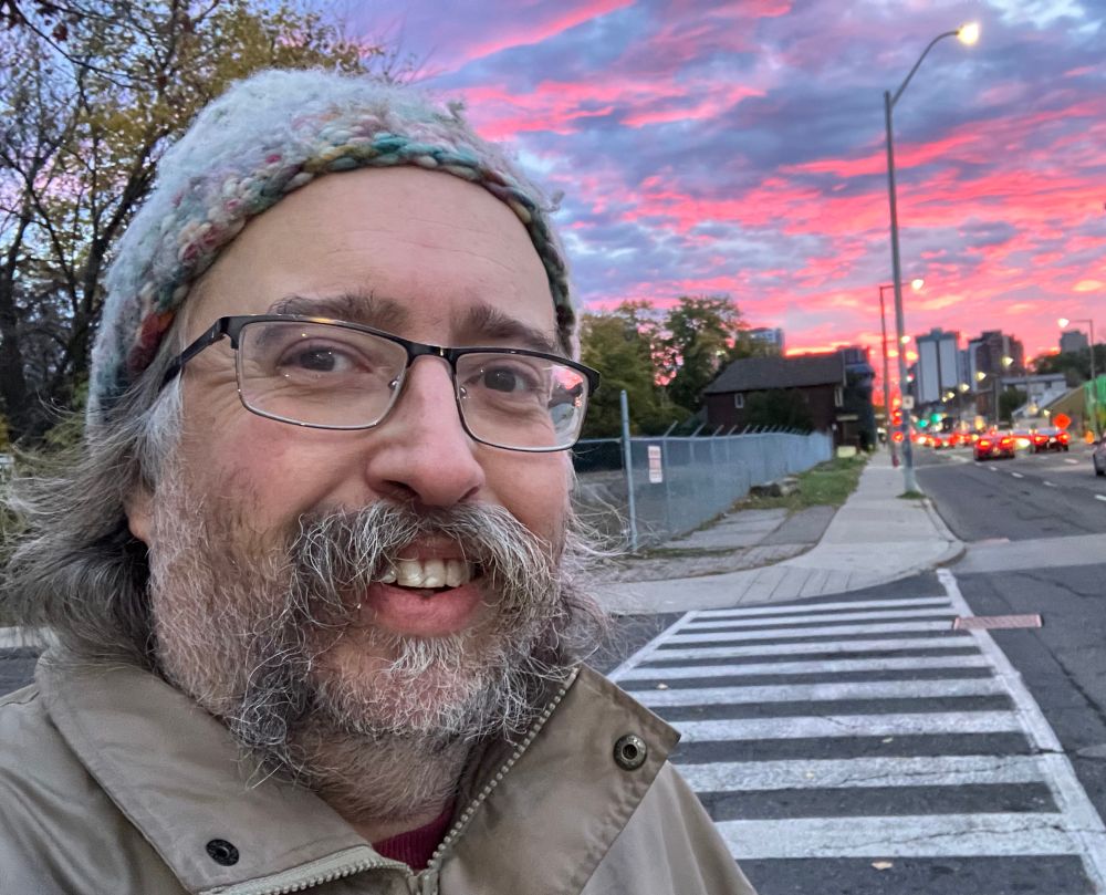 I am standing on Main Street in Hamilton (a white man of late middle age with grey-brown hair, stubble, a handlebar moustache, glasses and a beautiful multi pastel wool toque. The blue sky behind me is spattered with low-ish cloud blazing bright pink in the rising sun. Along the road cars' lights are glowing. 