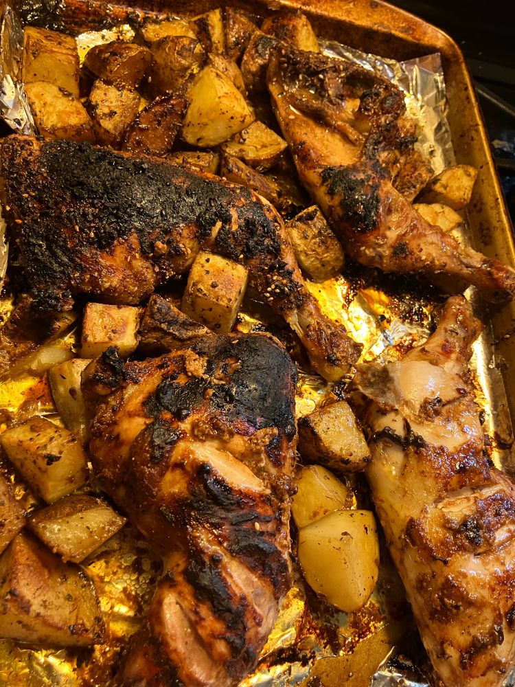 A sheet pan depicted in portrait, laden with broiled chicken leg quarters and chunks of baking potatoes. The leg quarters are tandoori style on the bottom of the frame and Korean sesame on the top. They are broiled dark brown with seared areas. The potatoes are herbed and scattered everywhere, dark in places having picked up all the chicken dripping
