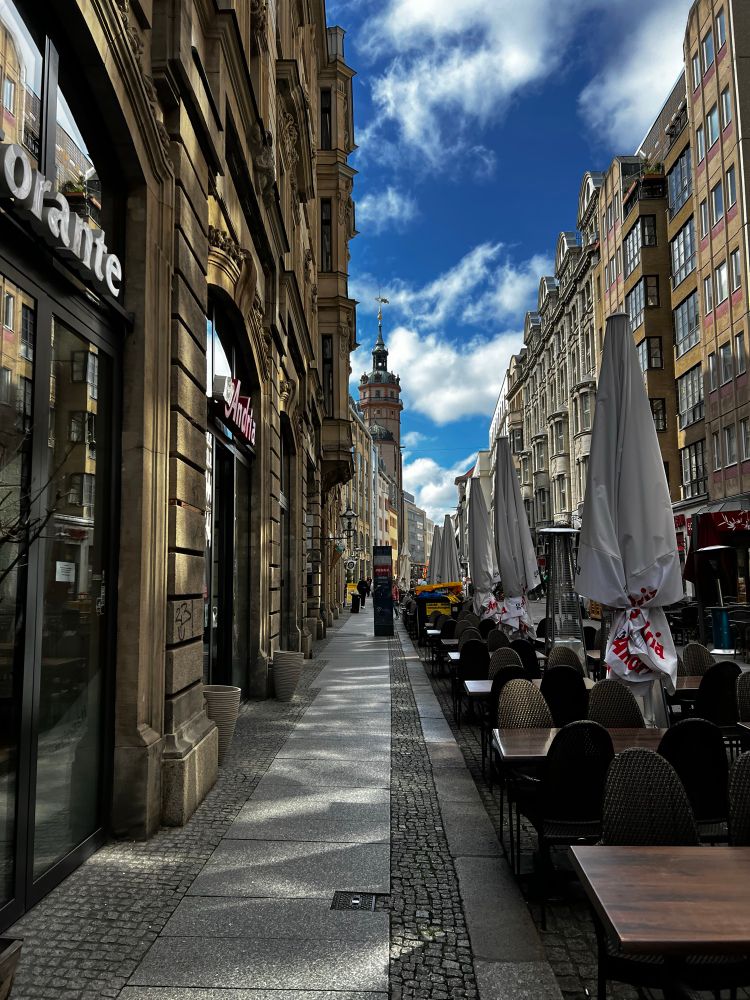 Nikolaistrasse in Leipzig, with cafe tables in the foreground and the Nikolaikirche tower just visible in the distance. It’s a sunny morning with a ble sky, the high buildings either side illuminated by the sun. 
