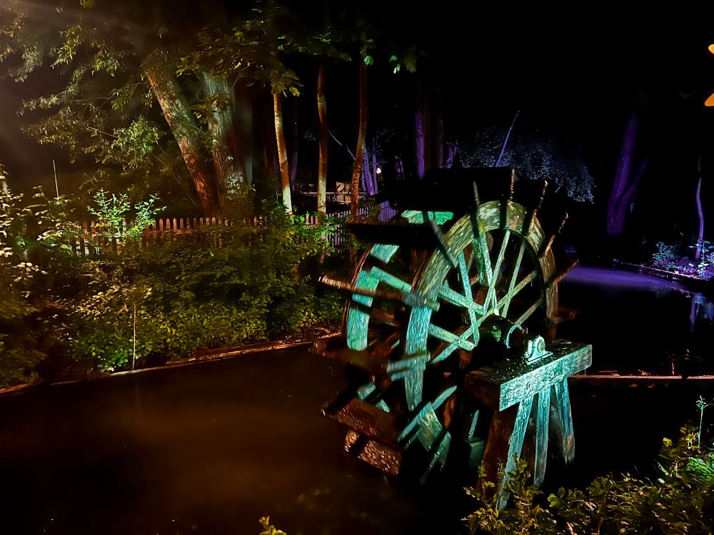 A nighttime photo of the wooden water wheel at Biergarten Mühlenpark, illuminated by coloured lights. 