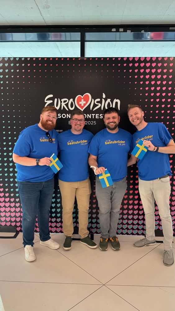 Ross, Trent, Tim, and Sean in front of a Eurovision banner. We have matching shirts in Swedish colors that say “vi är bastubröder nå jaa.”