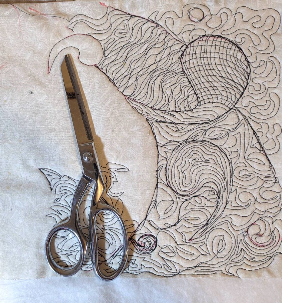 The image shows a pair of Gingher G-8 knife edge chrome sewing scissors placed on a piece of fabric with a complex, swirling stitched design. The scissors are positioned on the left side of the frame, lying diagonally with the blades pointing upwards and slightly to the right. The fabric design, occupying the right side and extending partially behind the scissors, features dense, meandering lines in dark thread, creating a textured, almost topographical effect. The fabric is a light, off-white color, providing a neutral backdrop that highlights the contrast of the stitching and the metallic sheen of the scissors. The overall composition suggests a focus on the tools and techniques of textile art, with the scissors serving as a prominent element against the backdrop of the stitched design.