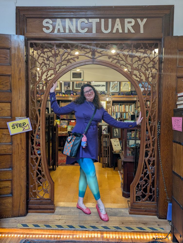 White woman wearing glasses and a very cool tunic/leggings outfit stands in a beautiful carved doorway that reads SANCTUARY above it 