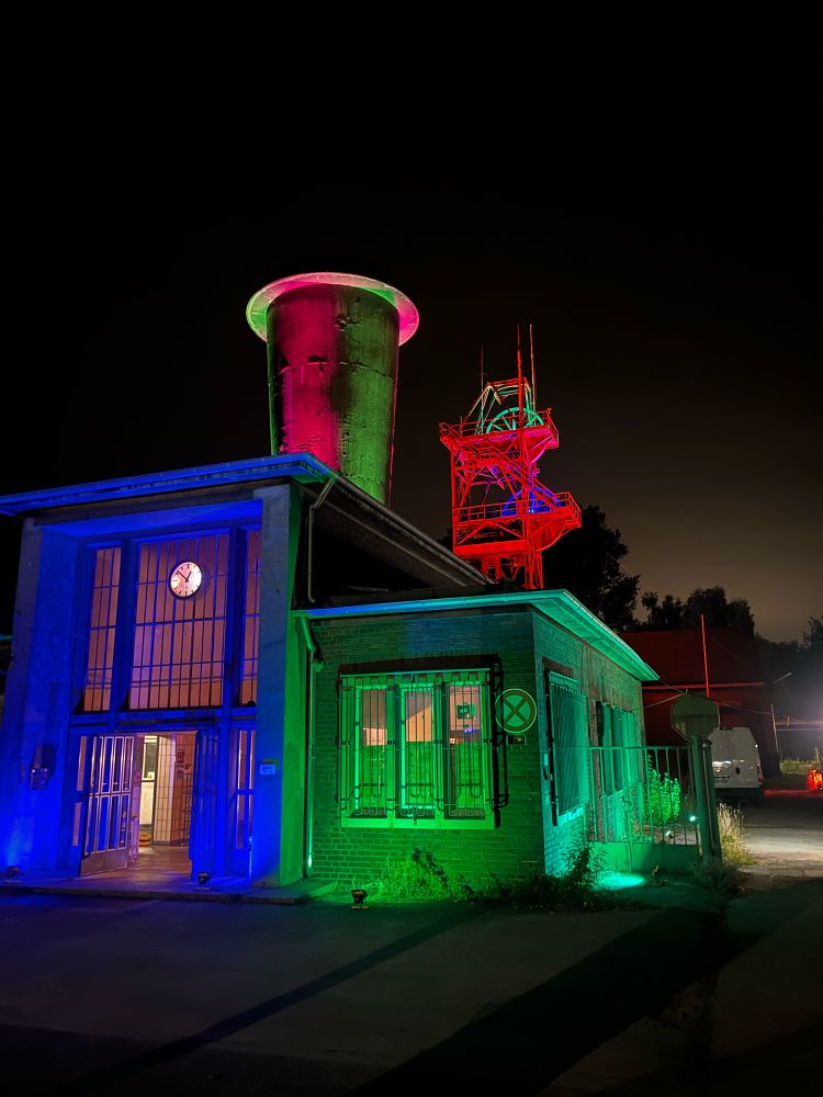Die Schachtanlage Blumenthal7 in Recklinghausen- zu sehen ist der Eingangsbereich, ein Grubenlüfter und das Fördergerüst - angestrahlt in blau und grün im Vordergrund sowie Pink-grün der Lüfter und rot mit grüner und blauer Seilscheibe das Fördergerüst