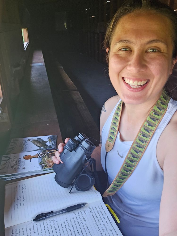 A selfie photo of a person with brown hair in a ponytail wearing a white tank top, sitting in a bird hide. They are holding binoculars with a green neck strap, and have a notebook and mini-guides in front of them on the small ledge