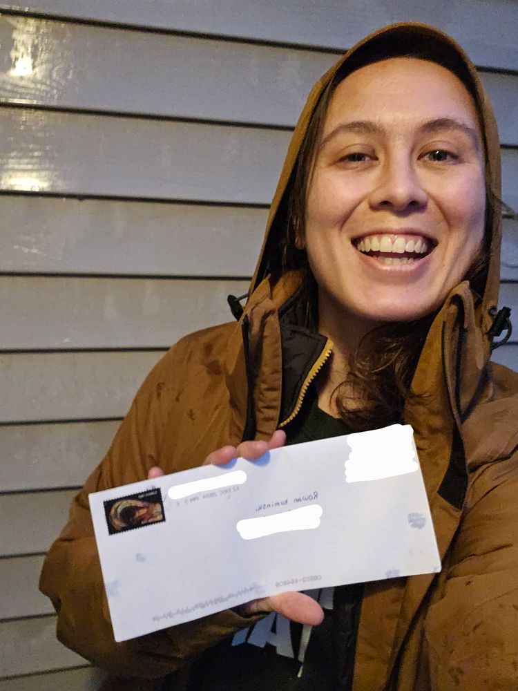 A selfie photo of a person with a brown winter coat with the hood up, as it's raining slightly. They're holding a white envelope and smiling