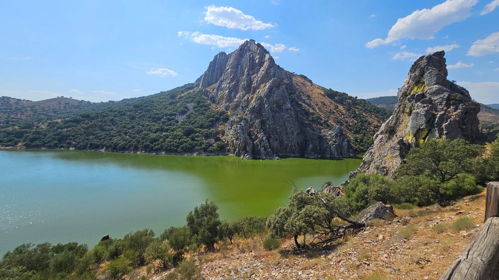 Photo of Monfragüe National Park. There are two rocky cliffs, one large one farther away in the center of the photo and a smaller one closer to the foreground. In between them is a green/blue body of water