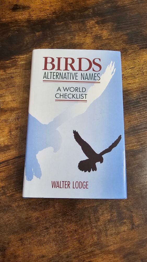 A photo of a book on a wooden surface: the book is a hardcover book with a white to sky blue gradient, and a black outline of a bird of prey flying in the bottom right of the cover, and the same outline but larger in blue on the left side of the book, extending to the back cover. The title says "Birds" in red, then in black letters the sub-title "Alternative Names: A World Checklist". The author's name, Walter Lodge, is written in red at the bottom of the book 