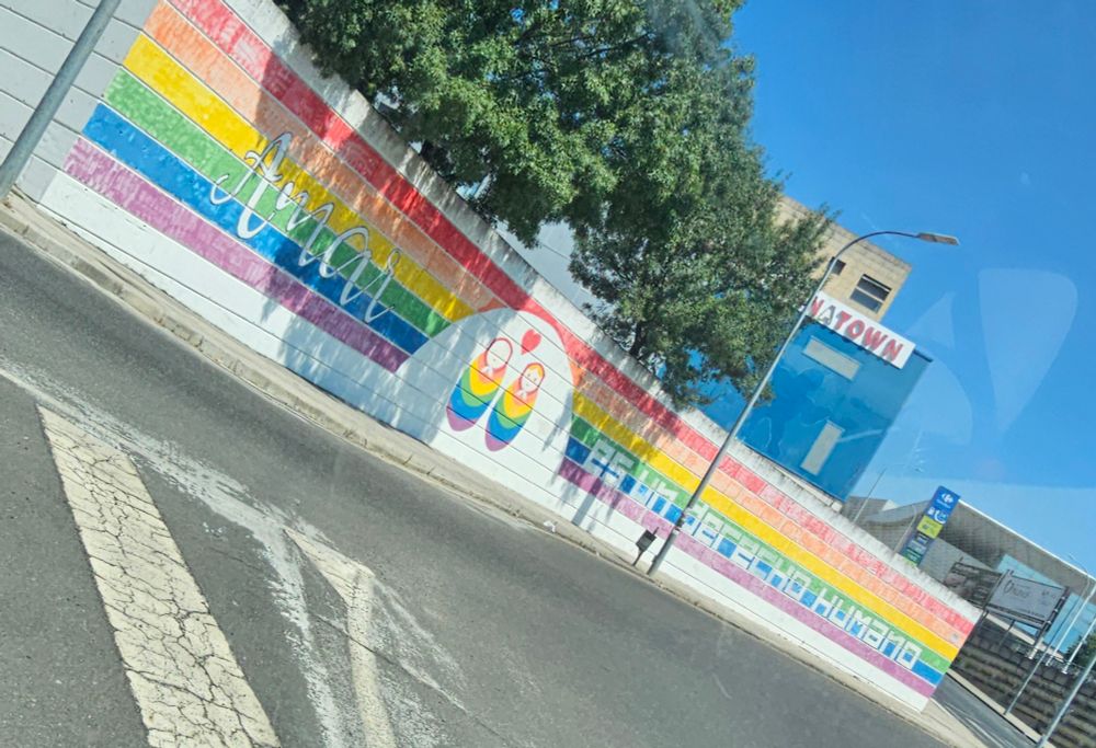 A photo of a painted wall at a roundabout. The wall is long and has a rainbow painted along it. In white cursive on the left is the word "Amar", and in white block letters after it says "es un derecho humano" (to love is a human right). In between the two blocks of text are two figurines in rainbow with a heart between them
