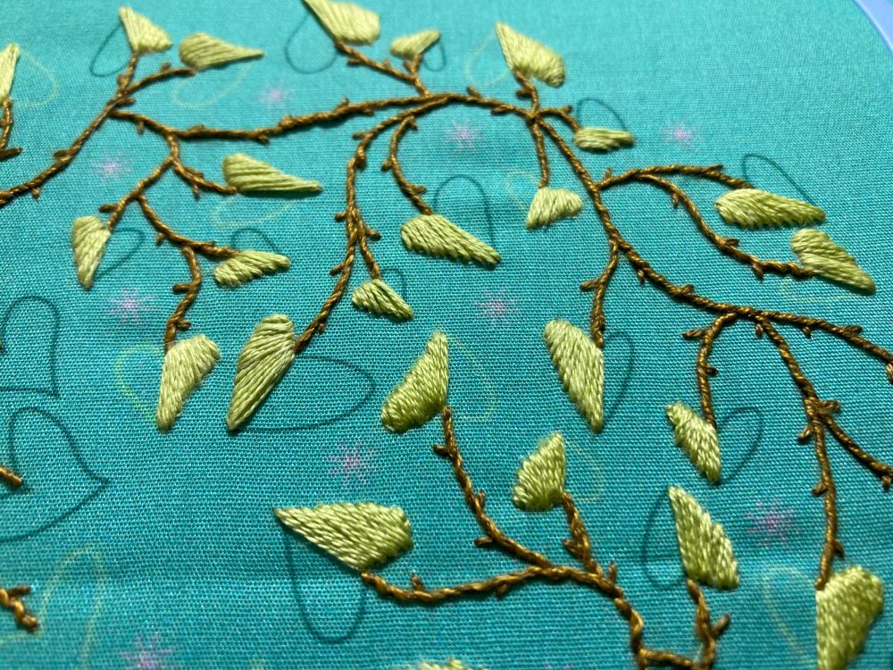 Embroidery pattern by Cozy Blue: close up of embroidered branches and partially completed leaves on aqua fabric. 