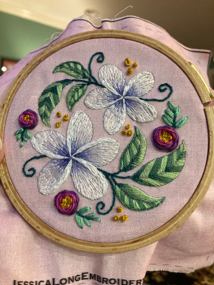 A completed Jessica Long embroidery pattern of two white and blue plumeria with wagon wheel roses and satin stitch leaves. 